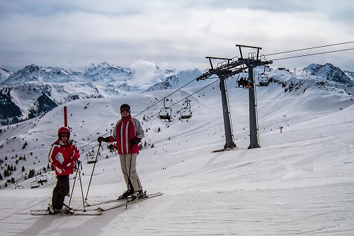 Two downhill skiers stand at the top of a snow covered mountain with a chair lift in the background